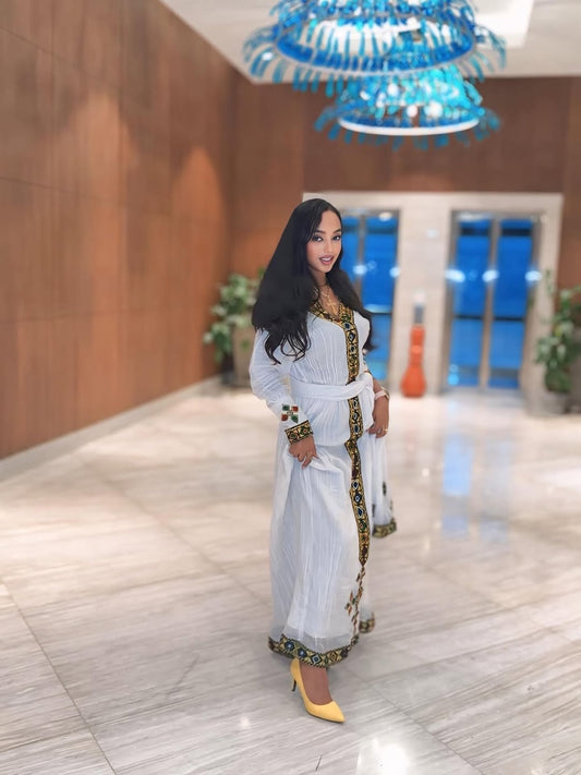 Woman in a white and gold habesha dress sitting on a decorative chair in an elegant interior setting.