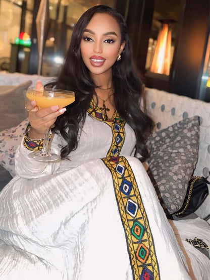 Woman in a white and gold habesha dress sitting on a decorative chair in an elegant interior setting.