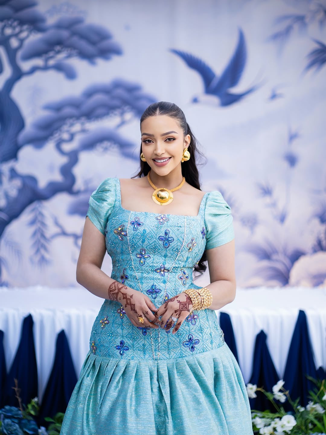Woman in a blue traditional Habasha dress   with floral patterns against a decorative wall.