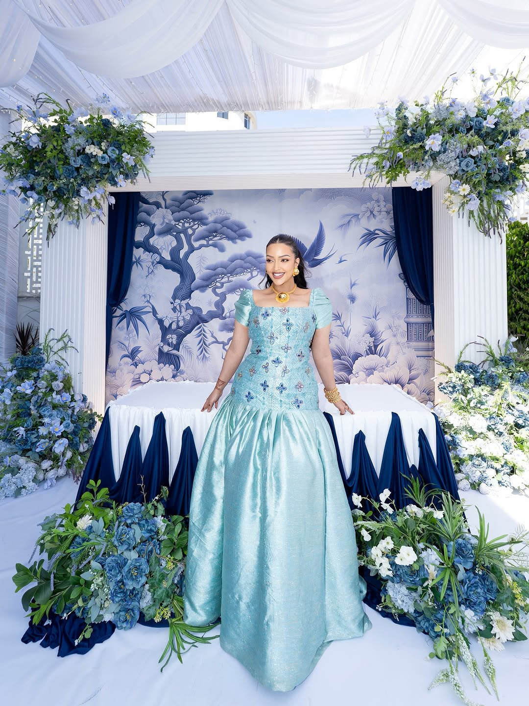 Woman in a light blue Habasha dress   standing in front of floral decorations and a decorated wall.