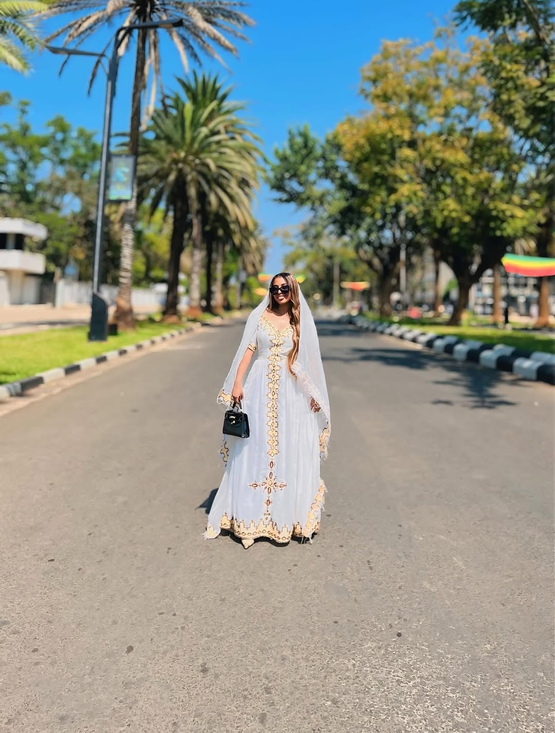 Woman in a white and gold habesha kemis standing on a road with palm trees in the background