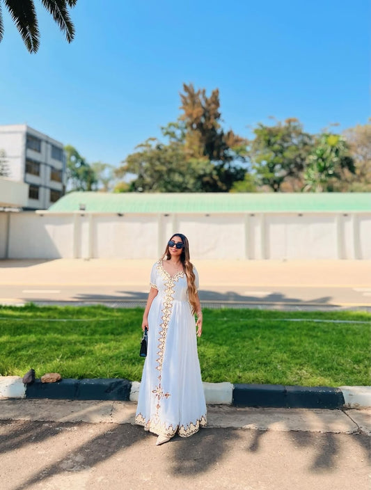 Woman in a white habesha kemis with gold patterns standing outdoors on a sunny day.