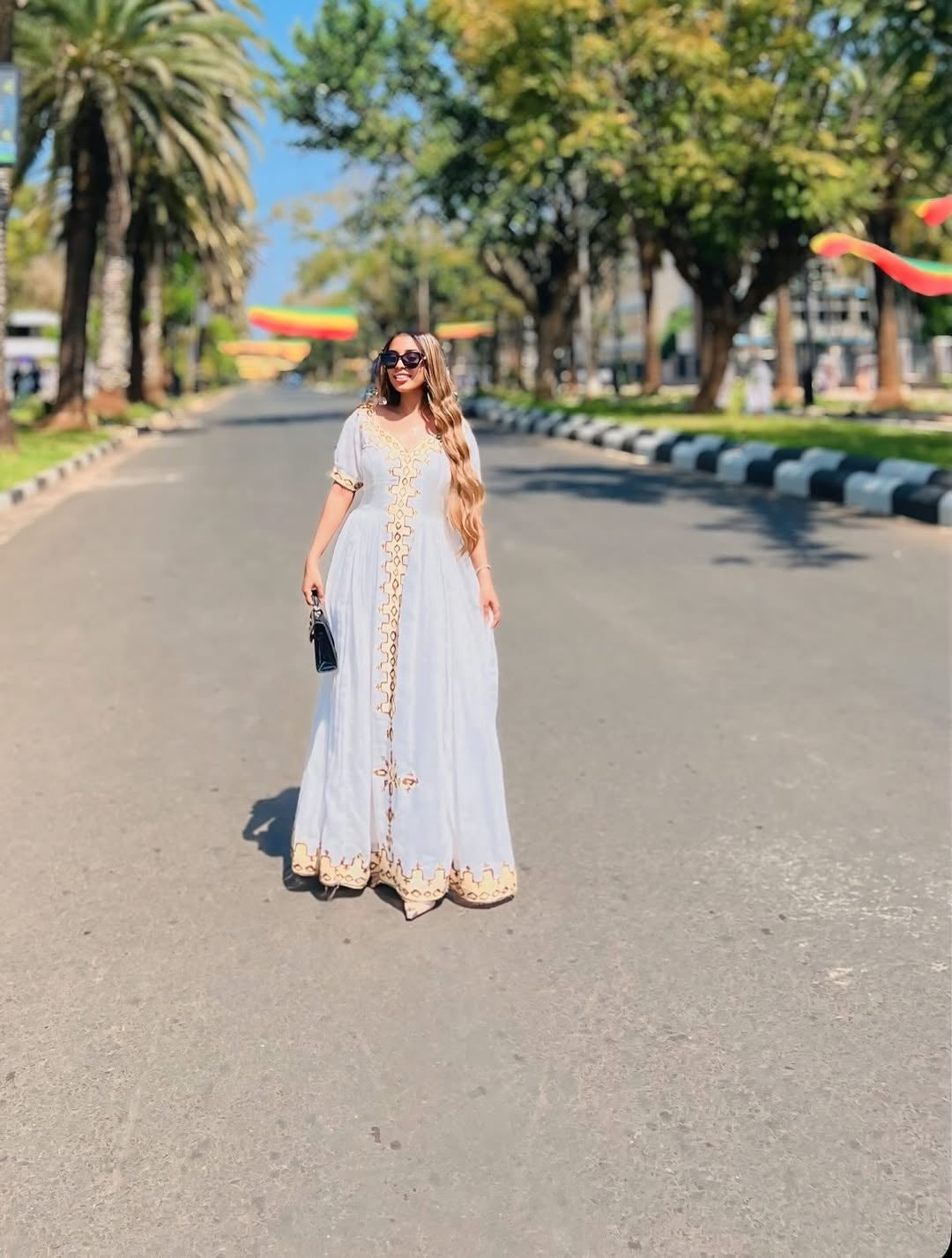 Woman in a long light-colored habesha kemis with gold accents standing on a tree-lined street.