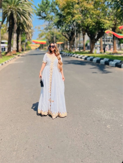 Woman in a long light-colored habesha kemis with gold accents standing on a tree-lined street.