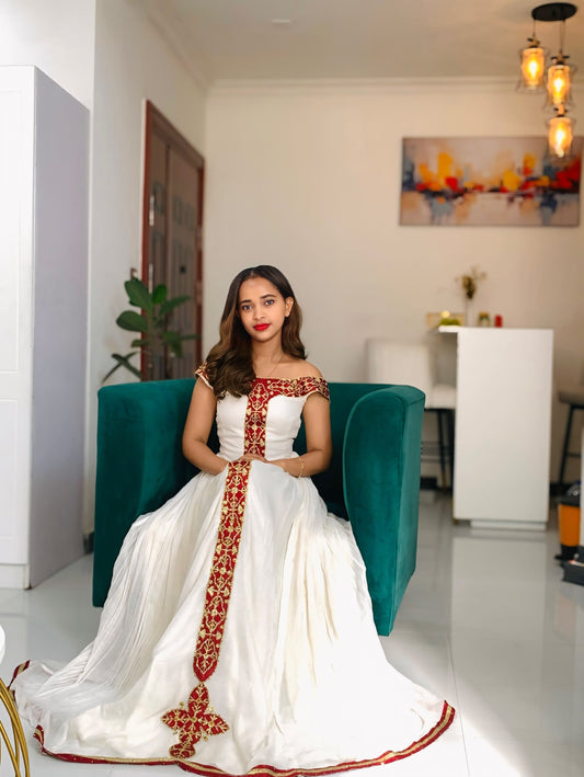 A woman wearing a white traditional Habesha dress  with red and gold embroidery sits on a teal armchair in a modern indoor setting.