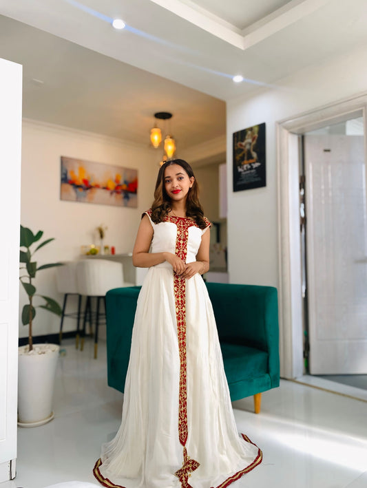 A woman wearing a white sleeveless maxi Habesha dress  with a vertical red and gold embroidered placket stands in a modern living room with a teal sofa and white walls.