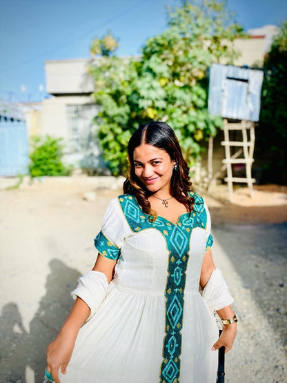 Woman in a white and teal traditional habesha dress standing outdoors with greenery in the background