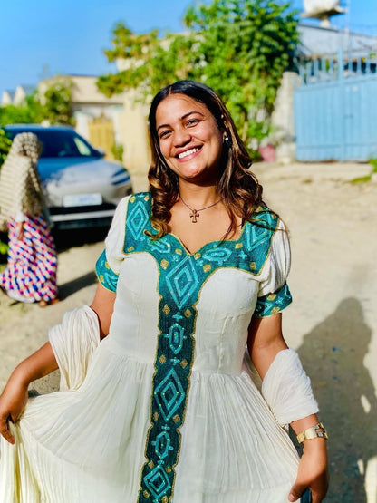 Woman in a white and green traditional Ethiopian kemis standing outdoors with a car and building in the background.