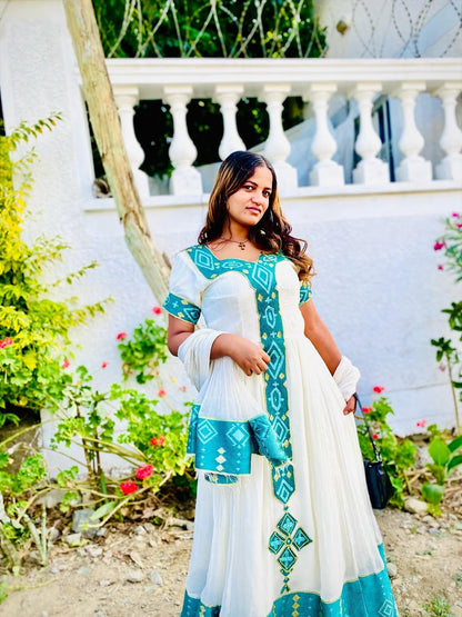 Woman in a white and blue traditional Ethiopian kemis standing outdoors with greenery and a white railing in the background.