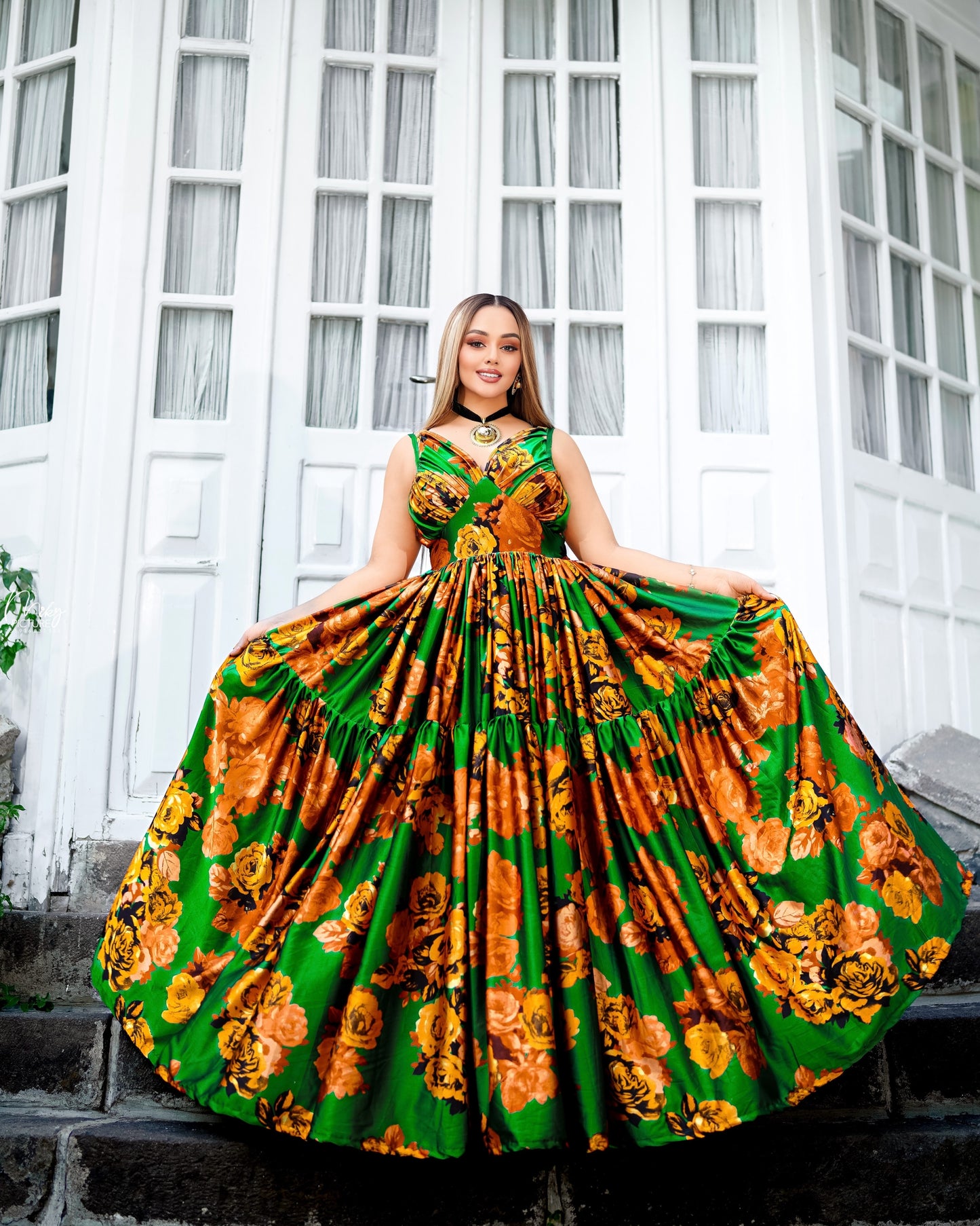 A woman stands on stone steps in front of a white building with tall window panes, wearing a voluminous sleeveless Habesha Chiffon with a green base and a bold orange and yellow floral print.