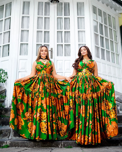 Two women wearing matching sleeveless A-line Habesha Chiffon with a vibrant green, orange, and yellow floral print, standing on stone steps in front of a white building with large window panes.