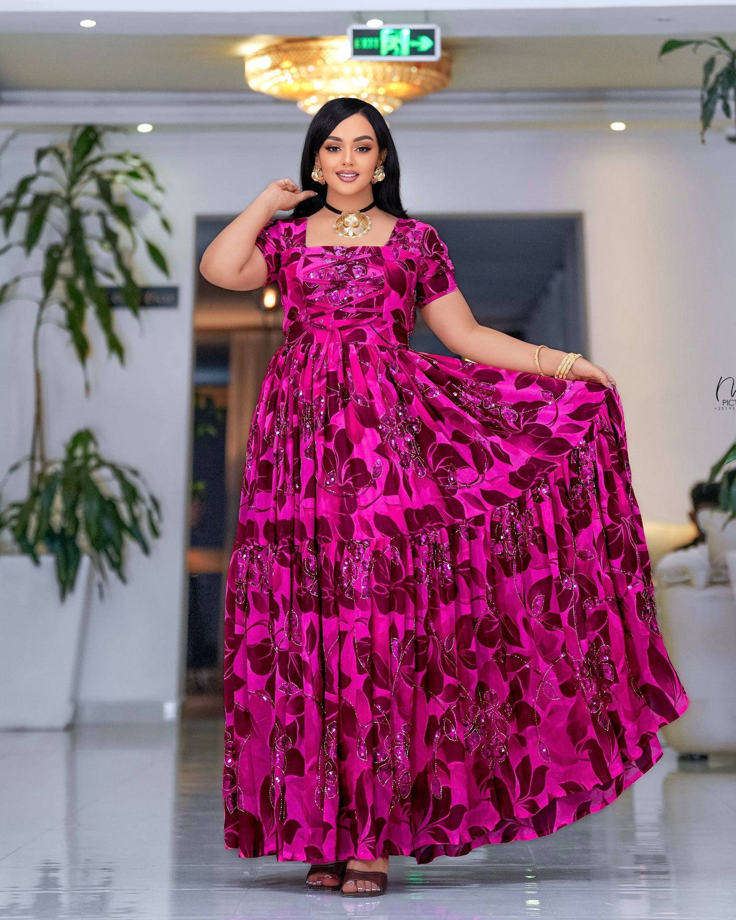 A woman modeling a vibrant  pink  Habesha dress with a dark red floral pattern, standing in a brightly lit indoor space.