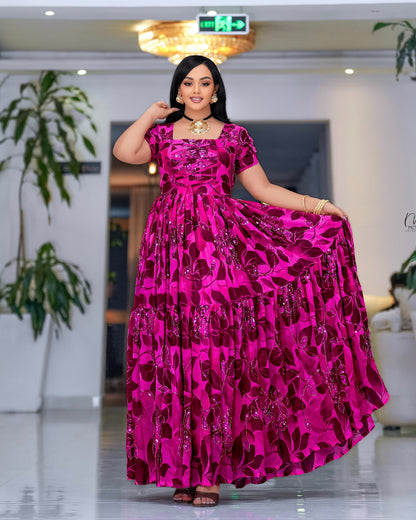 A woman modeling a vibrant  pink  Habesha dress with a dark red floral pattern, standing in a brightly lit indoor space.