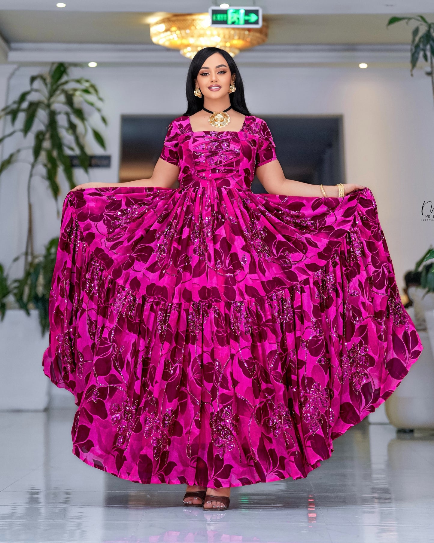 A woman modeling a bright magenta pink  Habesha dress  with a dark purple floral pattern, standing in a brightly lit indoor showroom.