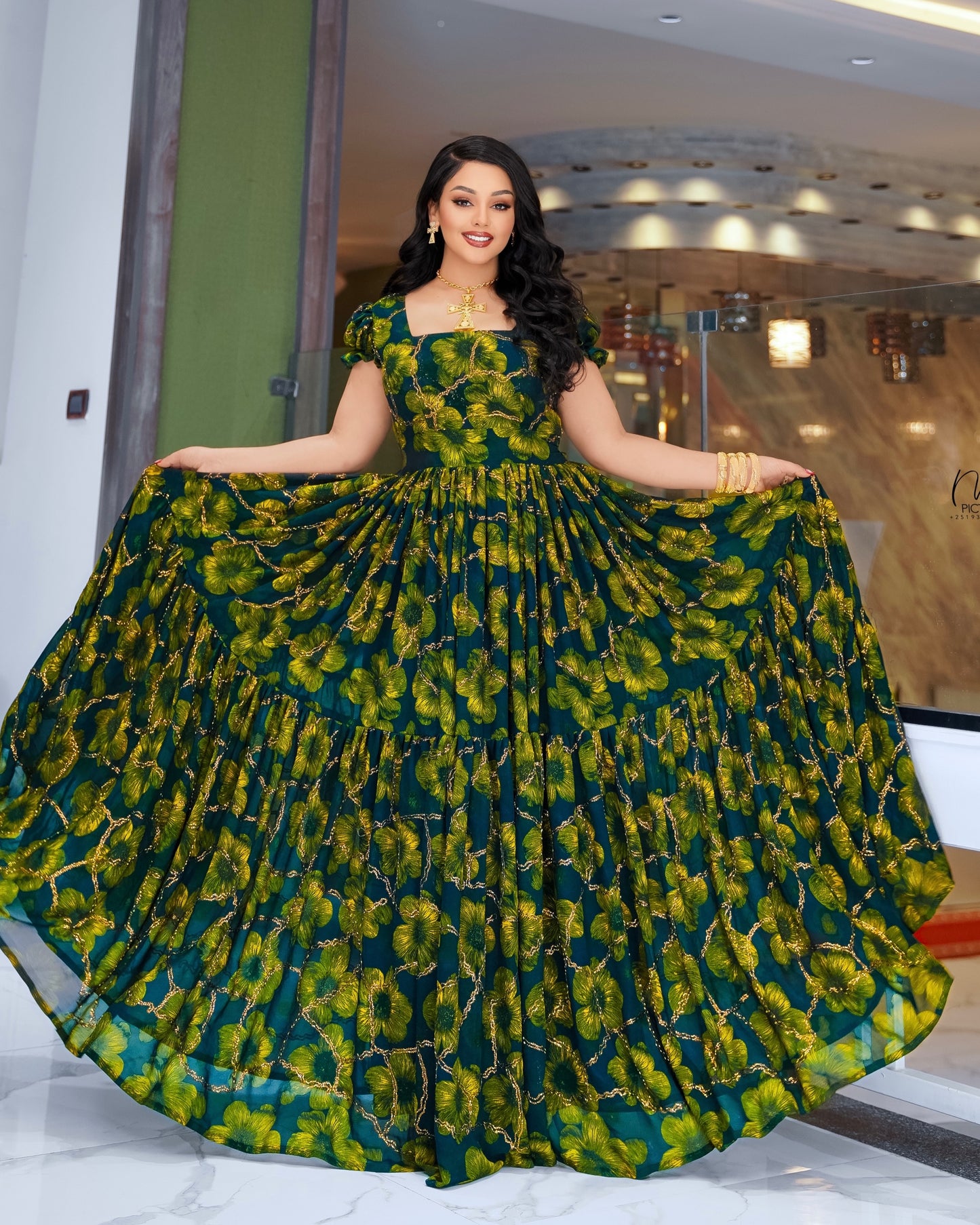 A woman modeling a floor-length green and yellow floral print Habesha Dress Chiffon with a tiered skirt, holding the sides of the skirt outwards to show its volume. The dress has a square neckline and short sleeves.