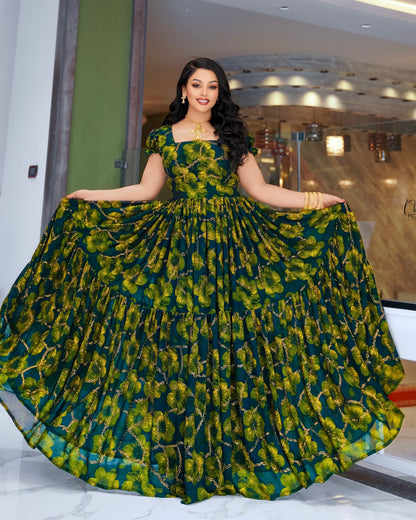A woman modeling a floor-length green and yellow floral print Habesha Dress Chiffon with a tiered skirt, holding the sides of the skirt outwards to show its volume. The dress has a square neckline and short sleeves.
