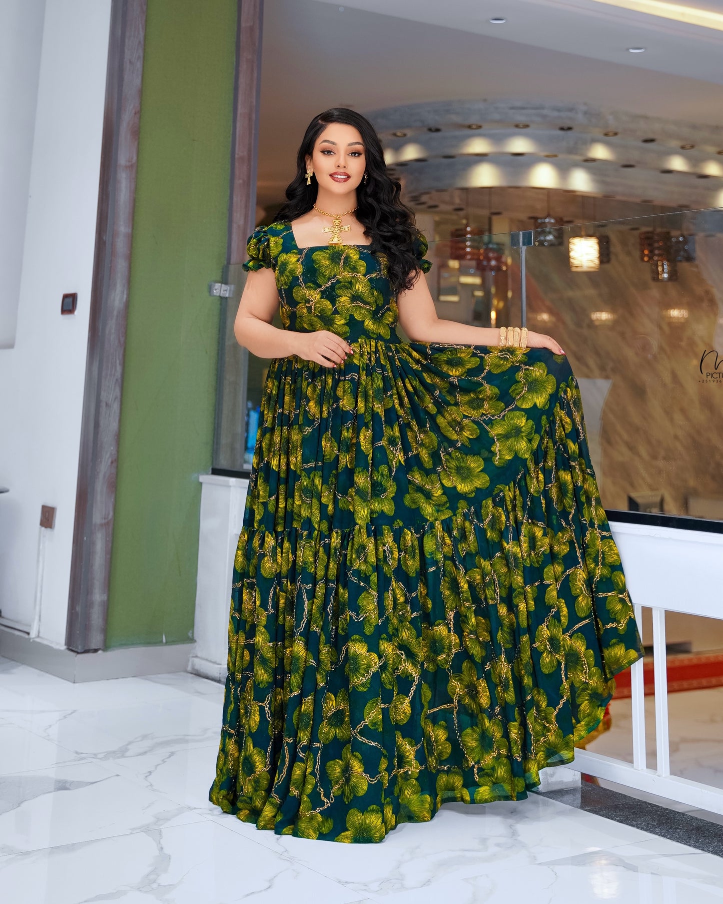 A woman modeling a floor-length, tiered Habesha Dress Chiffon with a green and yellow floral print, standing in a modern indoor setting with marble floors and glass walls.