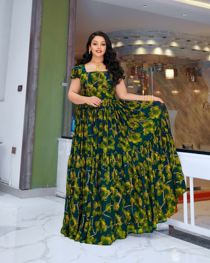 A woman modeling a floor-length, tiered Habesha Dress Chiffon with a green and yellow floral print, standing in a modern indoor setting with marble floors and glass walls.