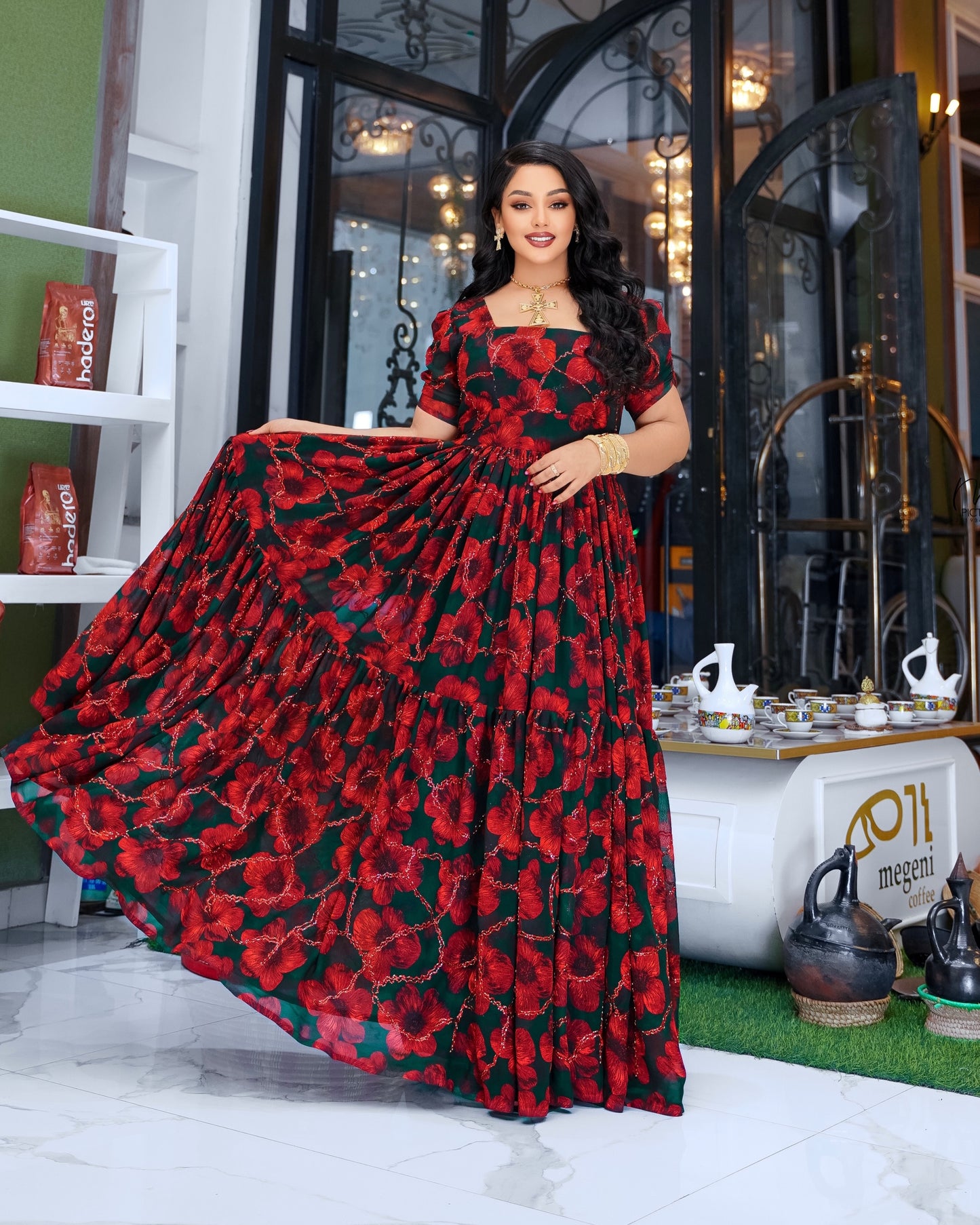 A woman models a floor-length tiered Habesha Chiffon  with a red and black floral print, holding out the skirt to show its volume in a decorated indoor setting.