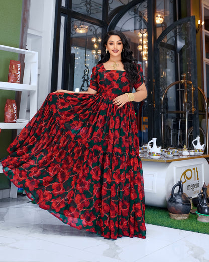 A woman models a floor-length tiered Habesha Chiffon  with a red and black floral print, holding out the skirt to show its volume in a decorated indoor setting.