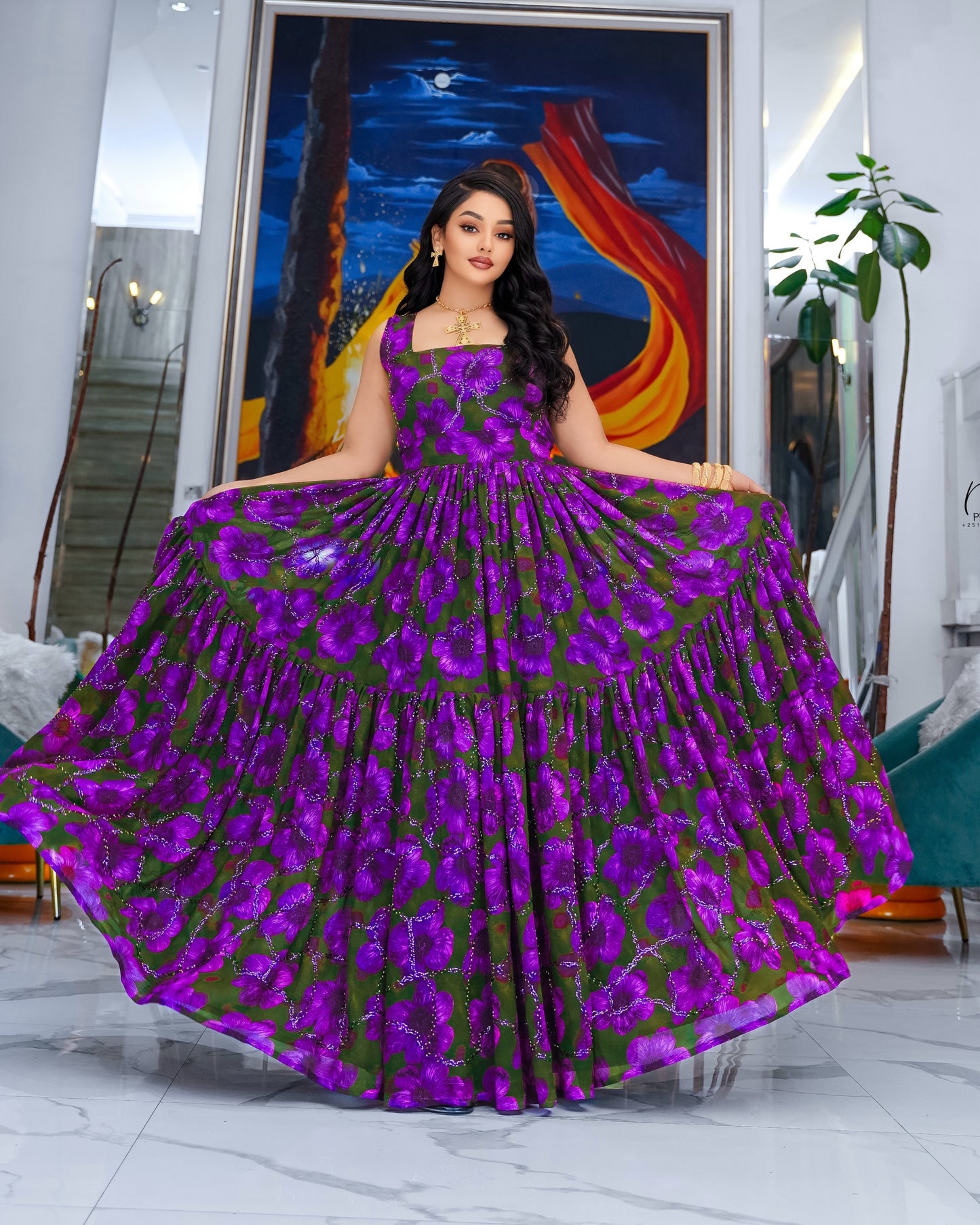 A woman stands indoors wearing a voluminous floor-length Habesha Chiffon  with a purple and green floral print. The dress has a fitted bodice and a very full tiered skirt, held out to show its scale.