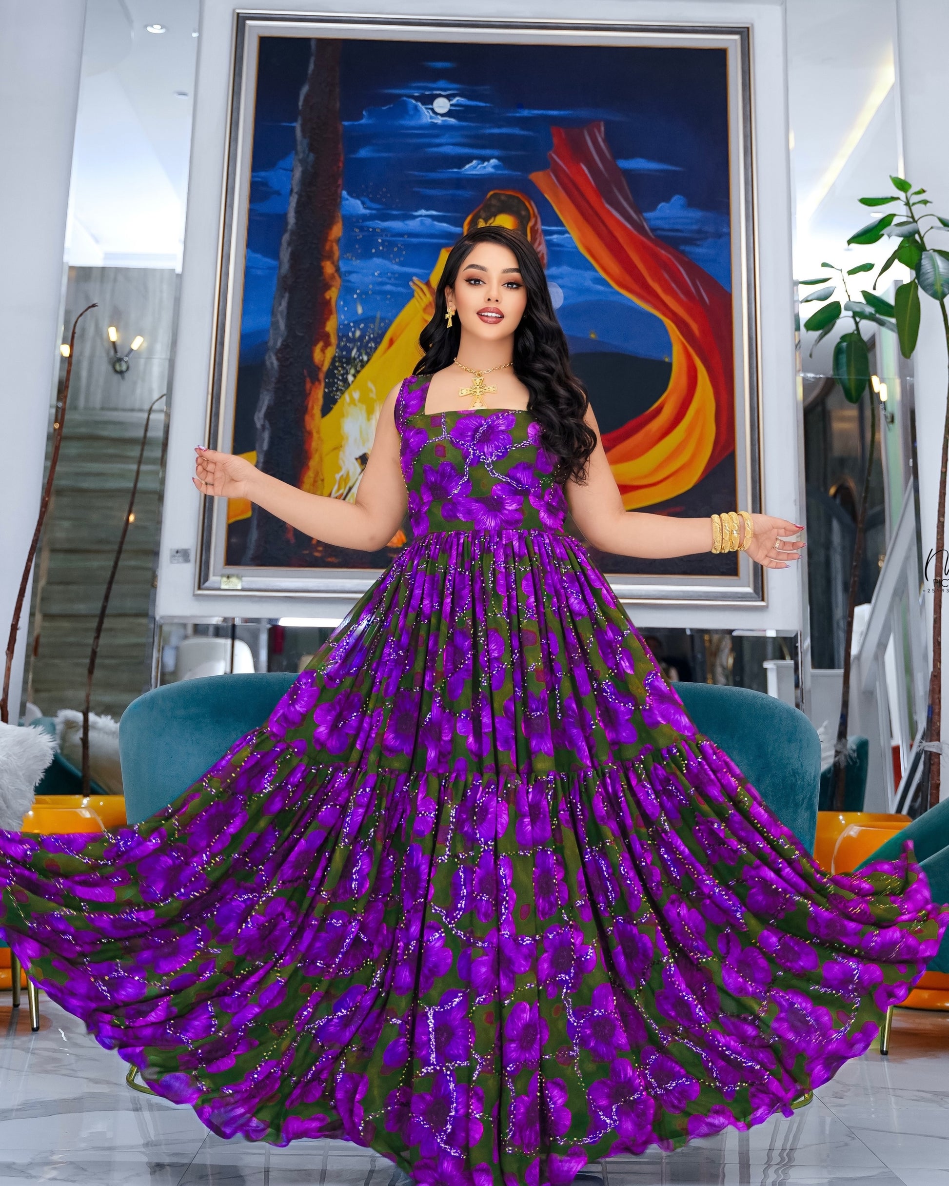 A woman stands in a room wearing a voluminous purple and green floral Habesha Chiffon  with sequins, holding out her arms to show the full skirt. A large framed painting hangs on the wall behind her.