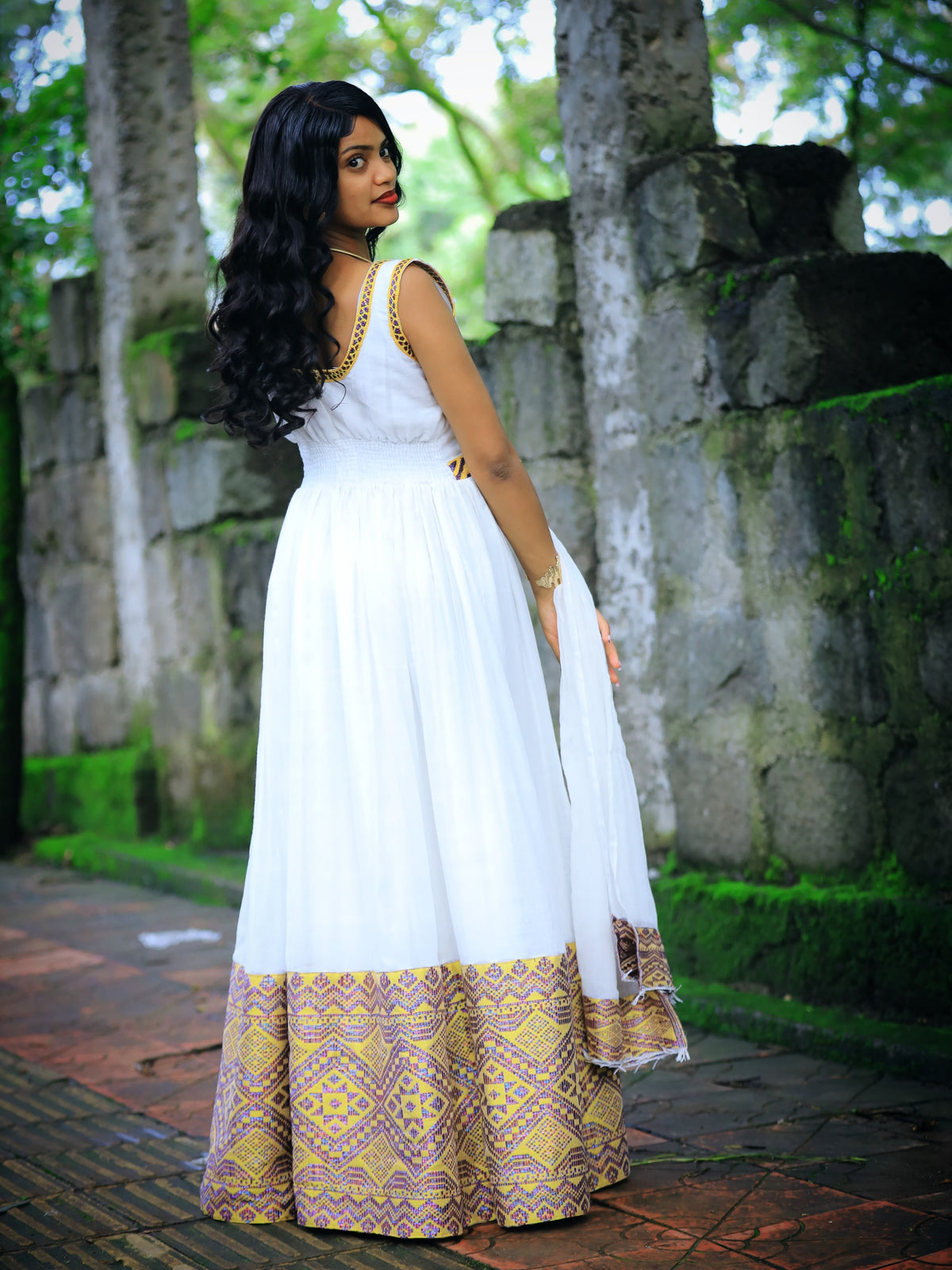 A woman wearing a white maxi Ethiopian Traditional Dress with gold trim and a gold and purple patterned border, standing outdoors in front of a stone wall and trees.