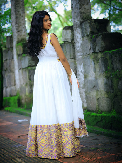 A woman wearing a white maxi Ethiopian Traditional Dress with gold trim and a gold and purple patterned border, standing outdoors in front of a stone wall and trees.