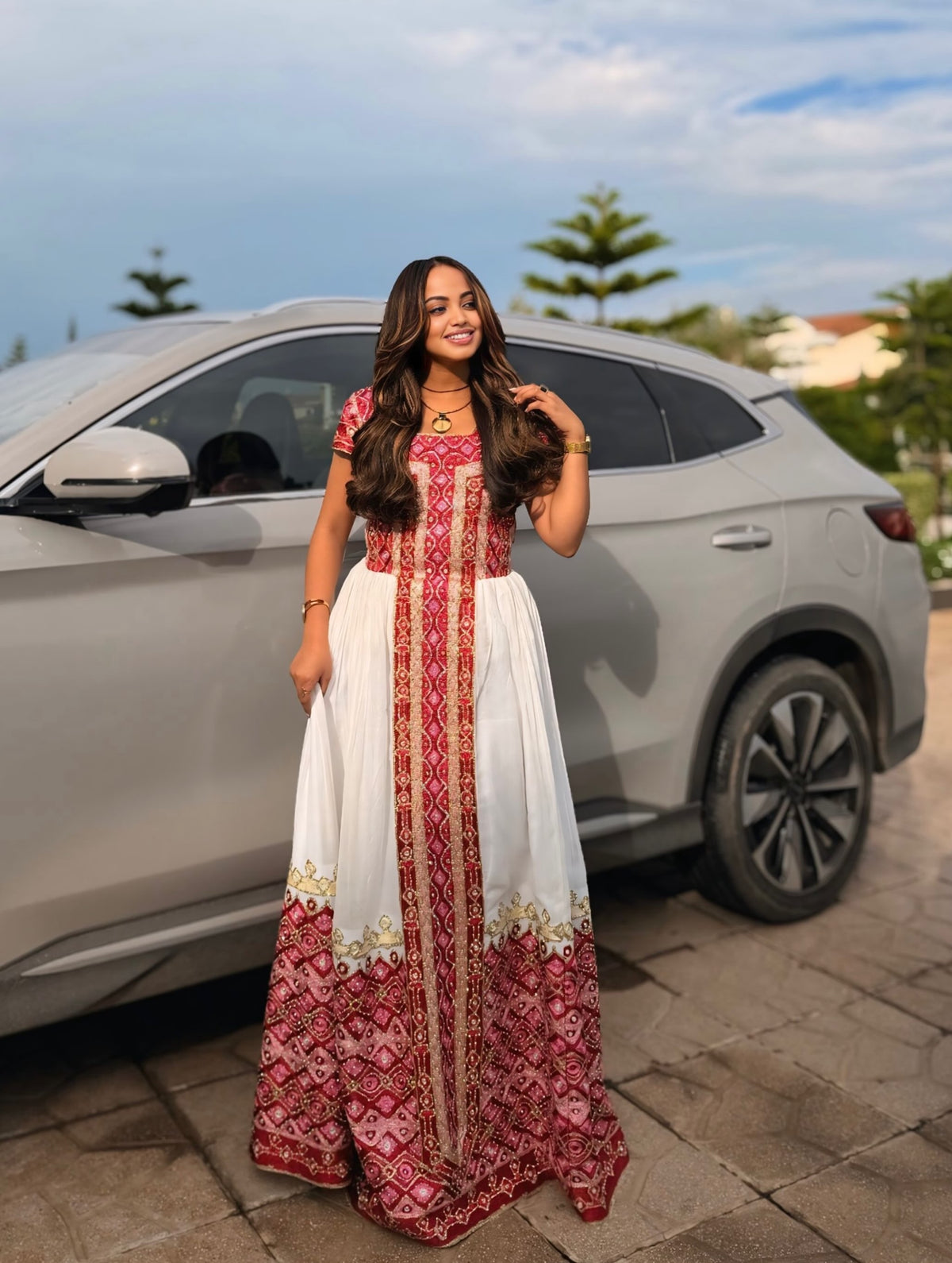 A woman wearing a long white and red embroidered Ethiopian clothing stands in front of a silver SUV outdoors on a paved area.