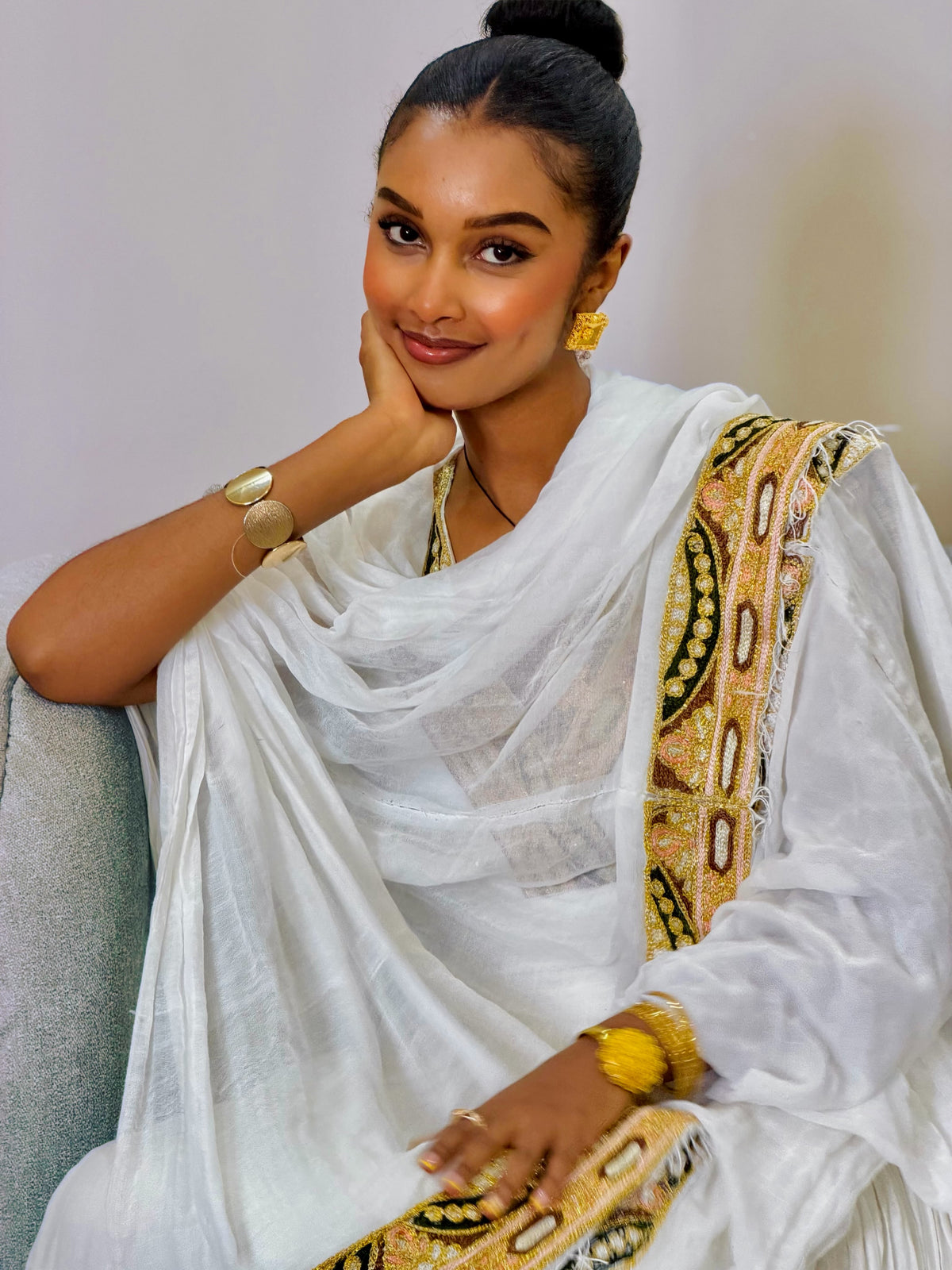 A woman wearing a white traditional draped Ethiopian Clothing with a gold and black patterned border, seated against a light gray sofa.
