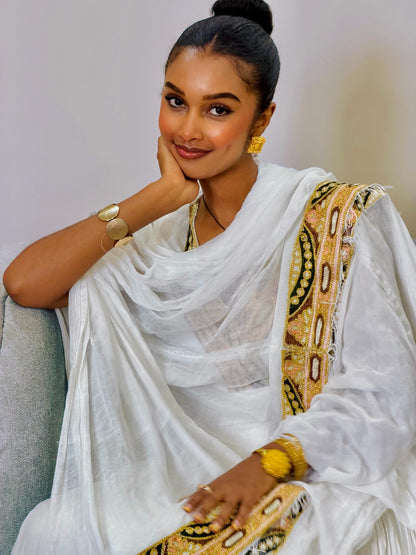A woman wearing a white traditional draped Ethiopian Clothing with a gold and black patterned border, seated against a light gray sofa.