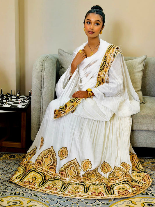 A woman seated on a gray sofa wearing a white traditional Ethiopian Clothing with ornate gold, beige, and brown embroidered patterns around the hem and along the draped fabric.