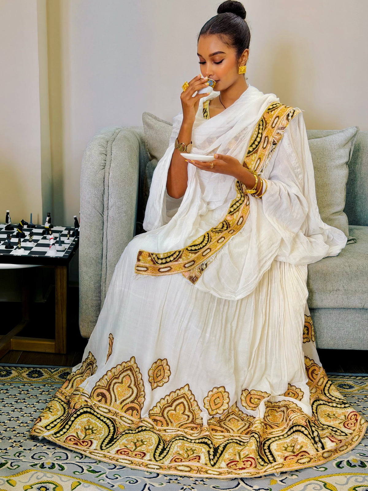 A woman with curly hair sits in a beige armchair, holding up a white and yellow patterned fabric Ethiopian Clothing in a modern living room setting.