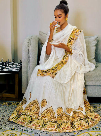 A woman with curly hair sits in a beige armchair, holding up a white and yellow patterned fabric Ethiopian Clothing in a modern living room setting.