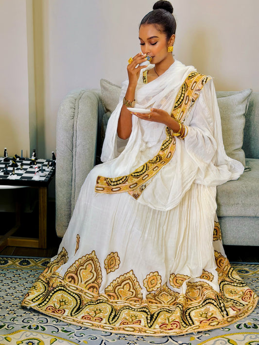 A woman with curly hair sits in a beige armchair, holding up a white and yellow patterned fabric Ethiopian Clothing in a modern living room setting.