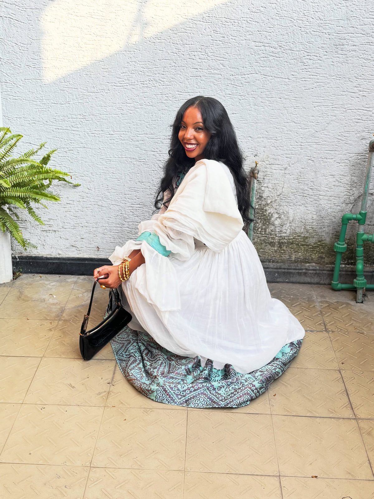 A woman sitting on a tiled patio against a textured white wall, wearing a white long-sleeve Habesha Kemis with a teal patterned hem and holding a black handbag.
