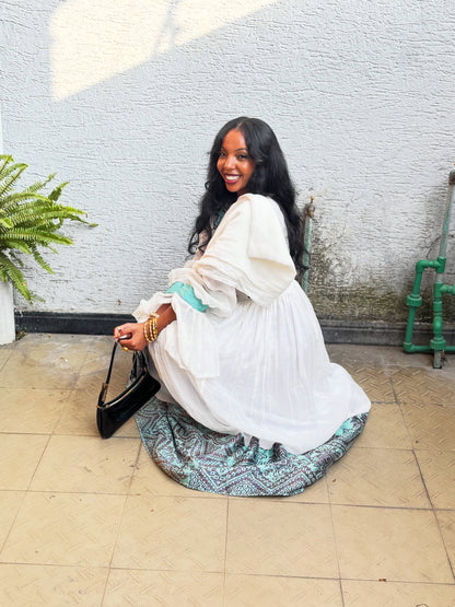 A woman sitting on a tiled patio against a textured white wall, wearing a white long-sleeve Habesha Kemis with a teal patterned hem and holding a black handbag.