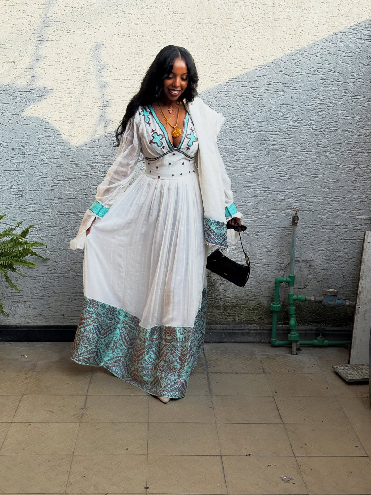 A woman wearing a white Habesha Kemis with turquoise and gold embroidery, a matching white shawl, and a patterned turquoise and gold hem, standing against a textured gray wall.