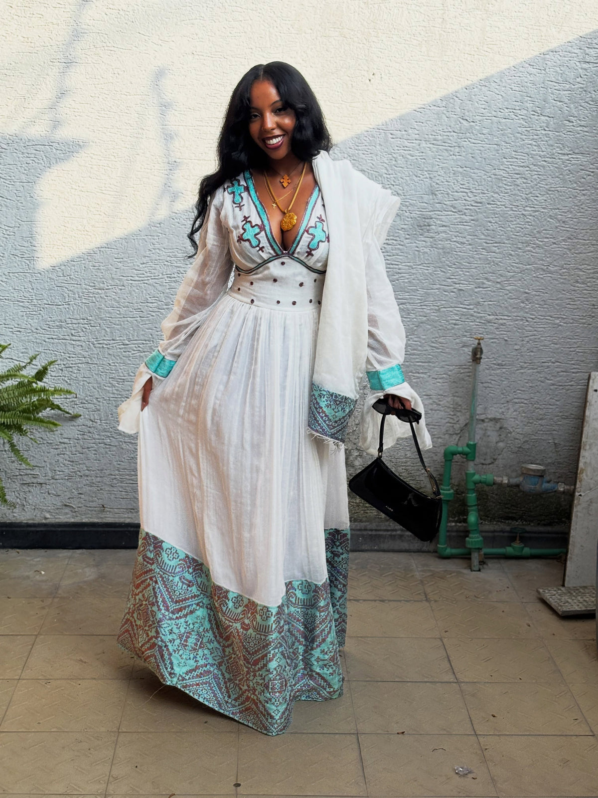 A woman wearing a white Habesha Kemis with turquoise and brown geometric patterned hem and cuffs, a matching embroidered bodice, and a white open-front cardigan, standing against a gray stucco wall.