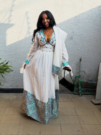 A woman wearing a white Habesha Kemis with turquoise and brown geometric patterned hem and cuffs, a matching embroidered bodice, and a white open-front cardigan, standing against a gray stucco wall.
