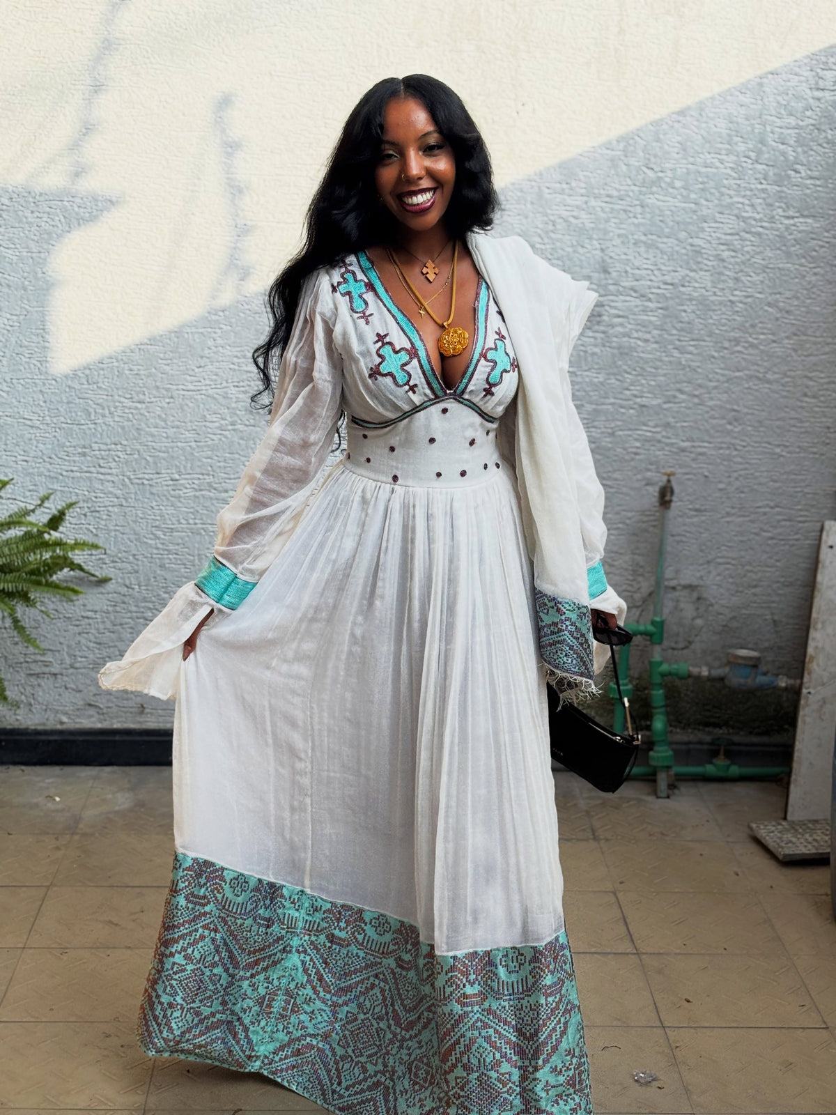 A woman wearing a white Habesha Kemis with turquoise and brown geometric patterns, a matching white open-front cardigan, and gold necklaces, standing against a textured gray wall.