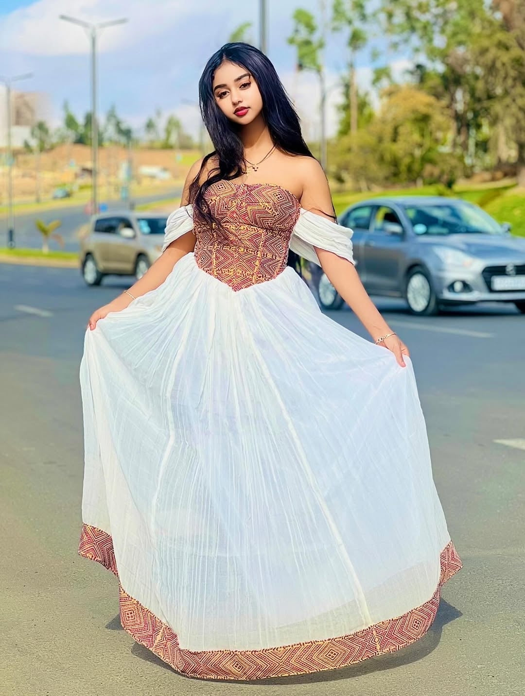 Woman in a white and patterned habesha dress standing on a road with cars and trees in the background