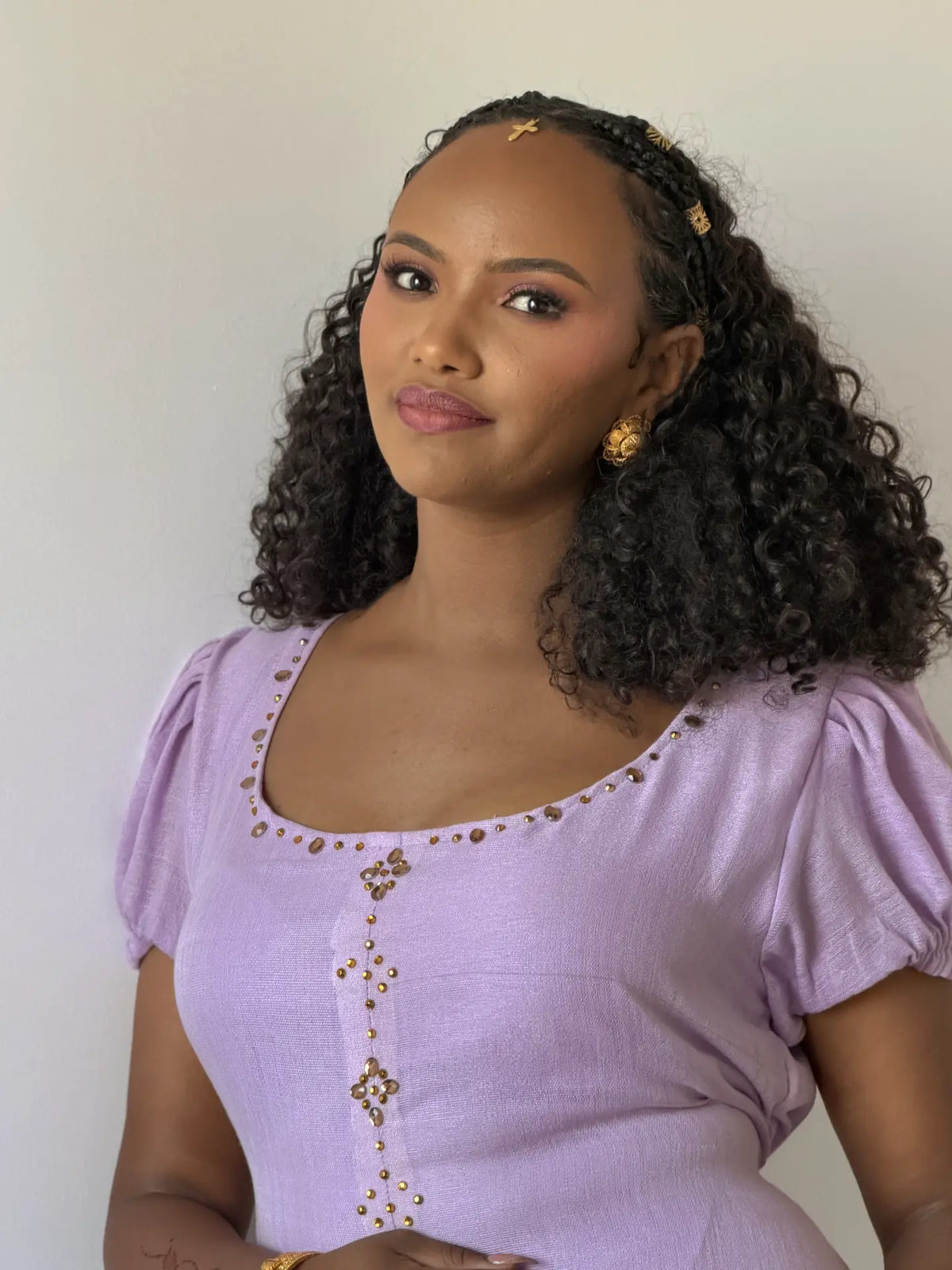 A woman with curly black hair wearing a lavender short-sleeve Ethiopian dress with gold embellishments, styled with a matching headband and gold earrings, against a plain off-white background.