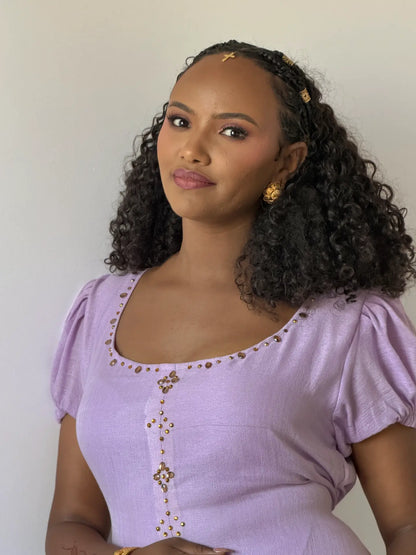 A woman with curly black hair wearing a lavender short-sleeve Ethiopian dress with gold embellishments, styled with a matching headband and gold earrings, against a plain off-white background.