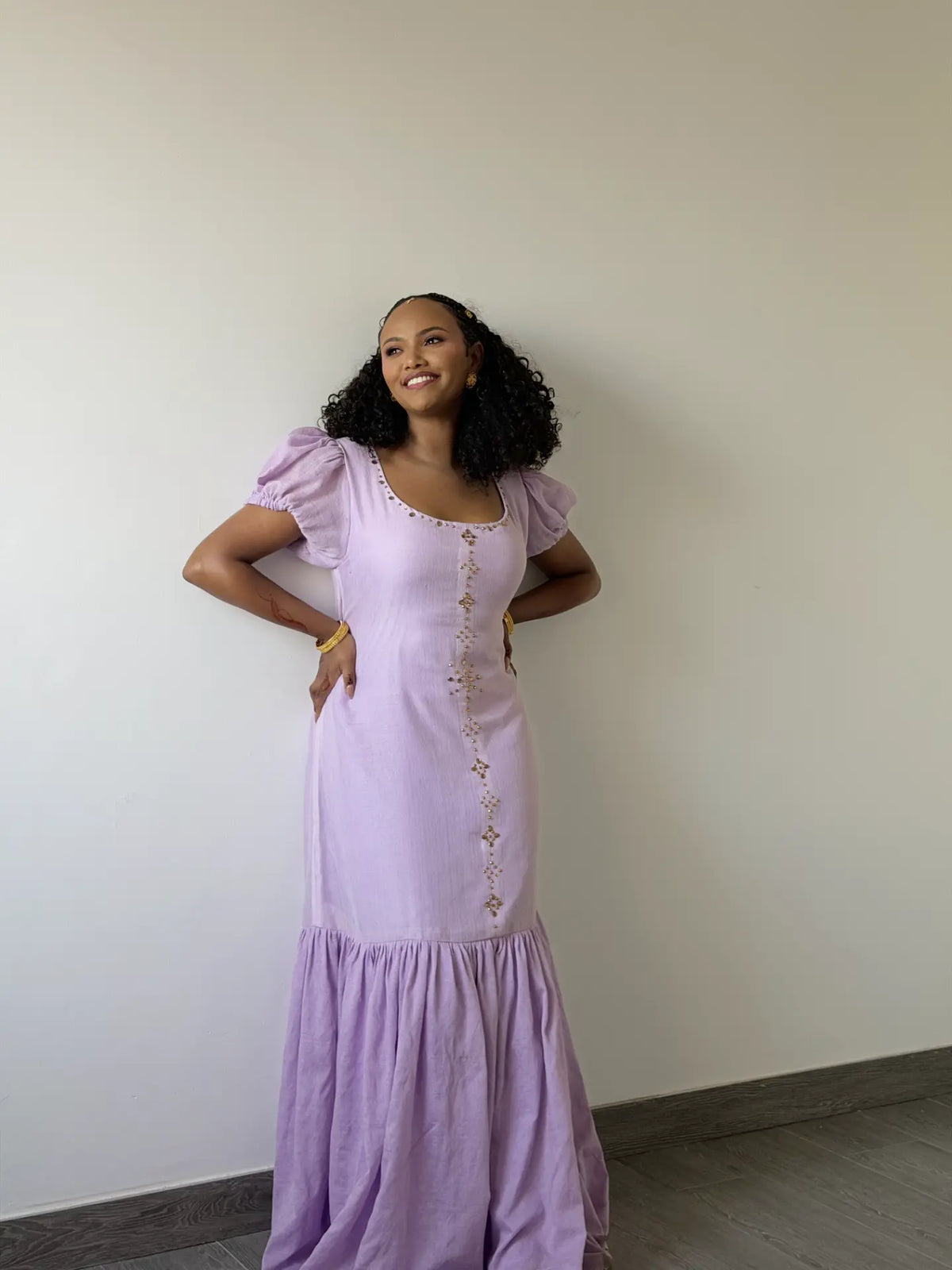 A woman wearing a lavender Ethiopian dress with puff sleeves and a decorative embroidered front, posing against a plain off-white wall.