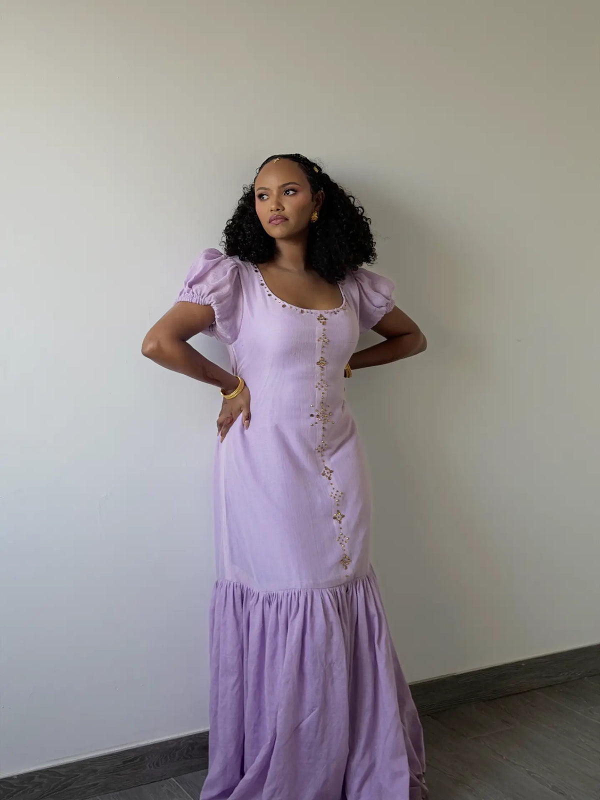 A woman wearing a lavender purple Ethiopian dress with gold metallic embellishments along the neckline and center front, styled with gold jewelry and henna on her hands.