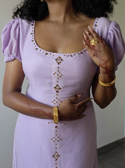 A woman wearing a lavender purple Ethiopian dress with gold metallic embellishments along the neckline and center front, styled with gold jewelry and henna on her hands.