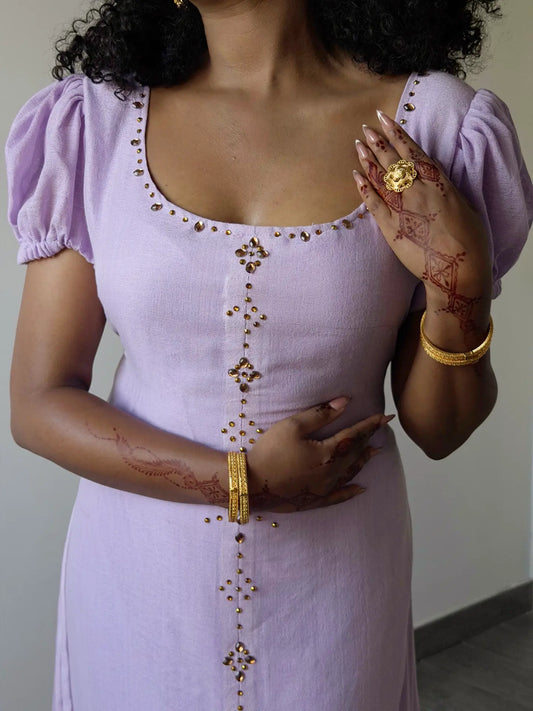 A woman wearing a lavender purple Ethiopian dress with gold metallic embellishments along the neckline and center front, styled with gold jewelry and henna on her hands.