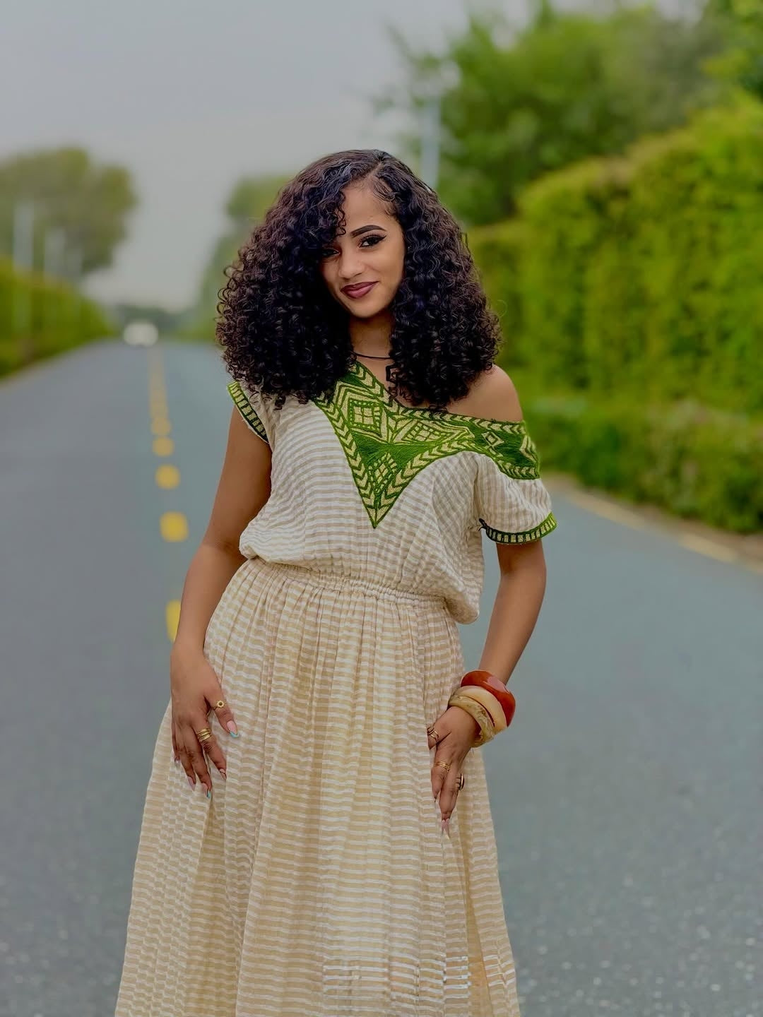 Woman in a patterned habesha dress standing on a road with greenery in the background