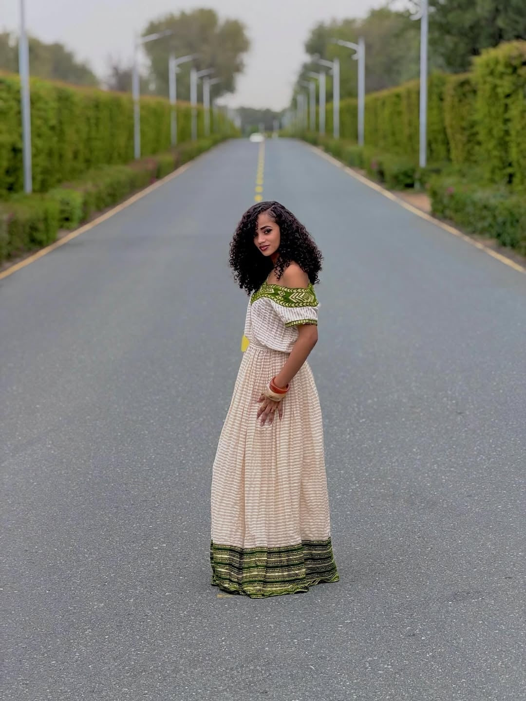 Woman in a green and beige habesha dress  standing on a road with greenery on either side.